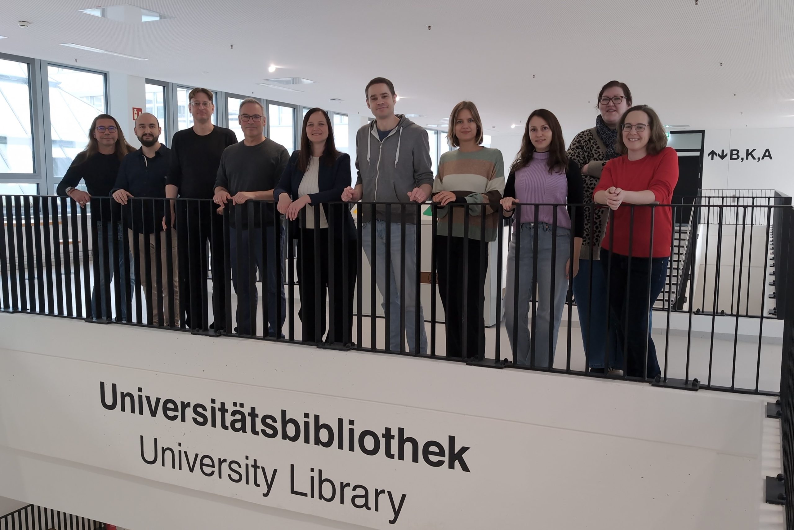 Die openCost Projektgruppe stehen nebeneinander an einem Geländer über dem Schriftzug "Universitätsbibliothek"
